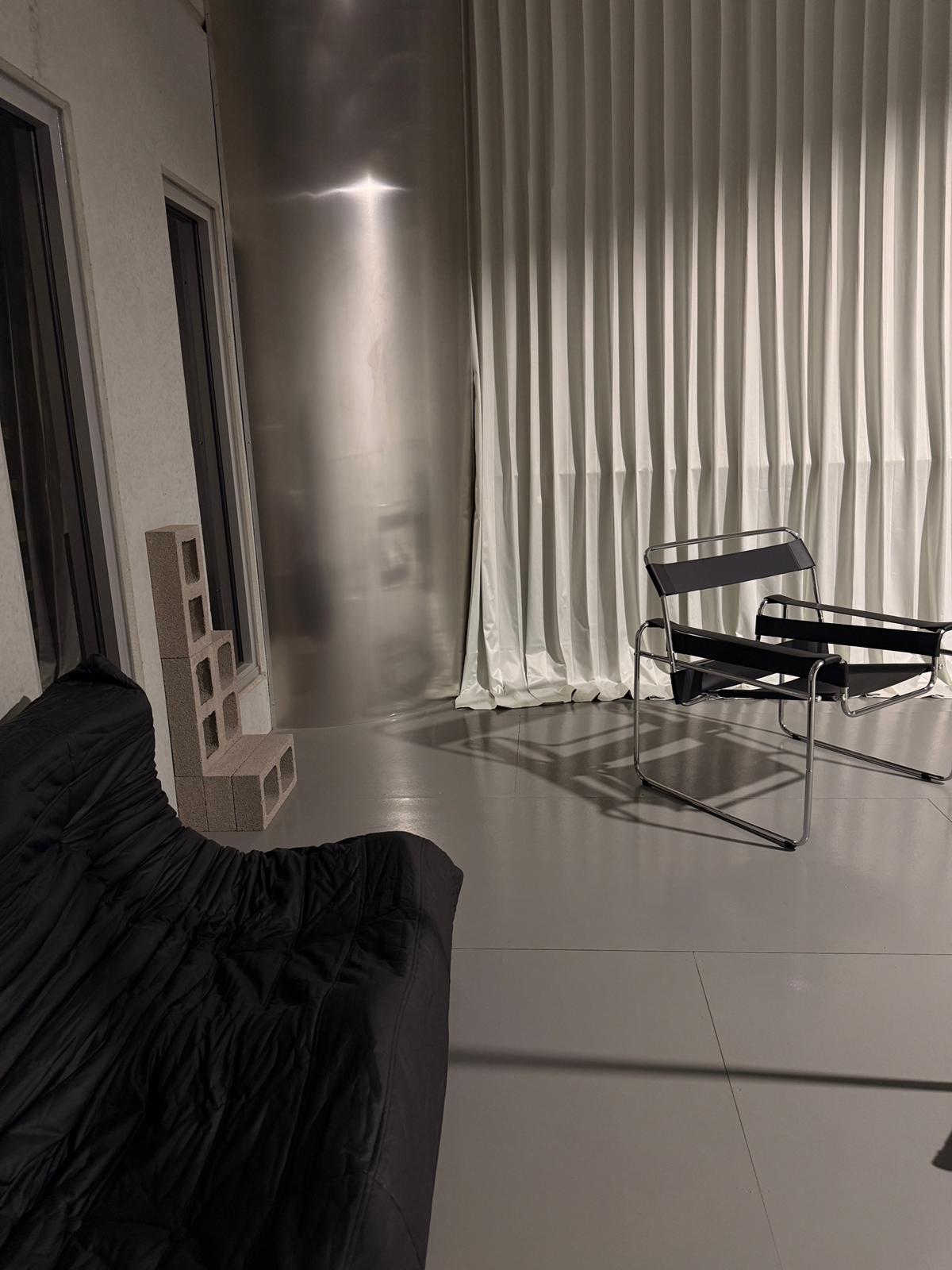 Night view of the studio with a Wassily chair, block-out curtains, breeze blocks and a black Togo sofa, lit by a single warm light.