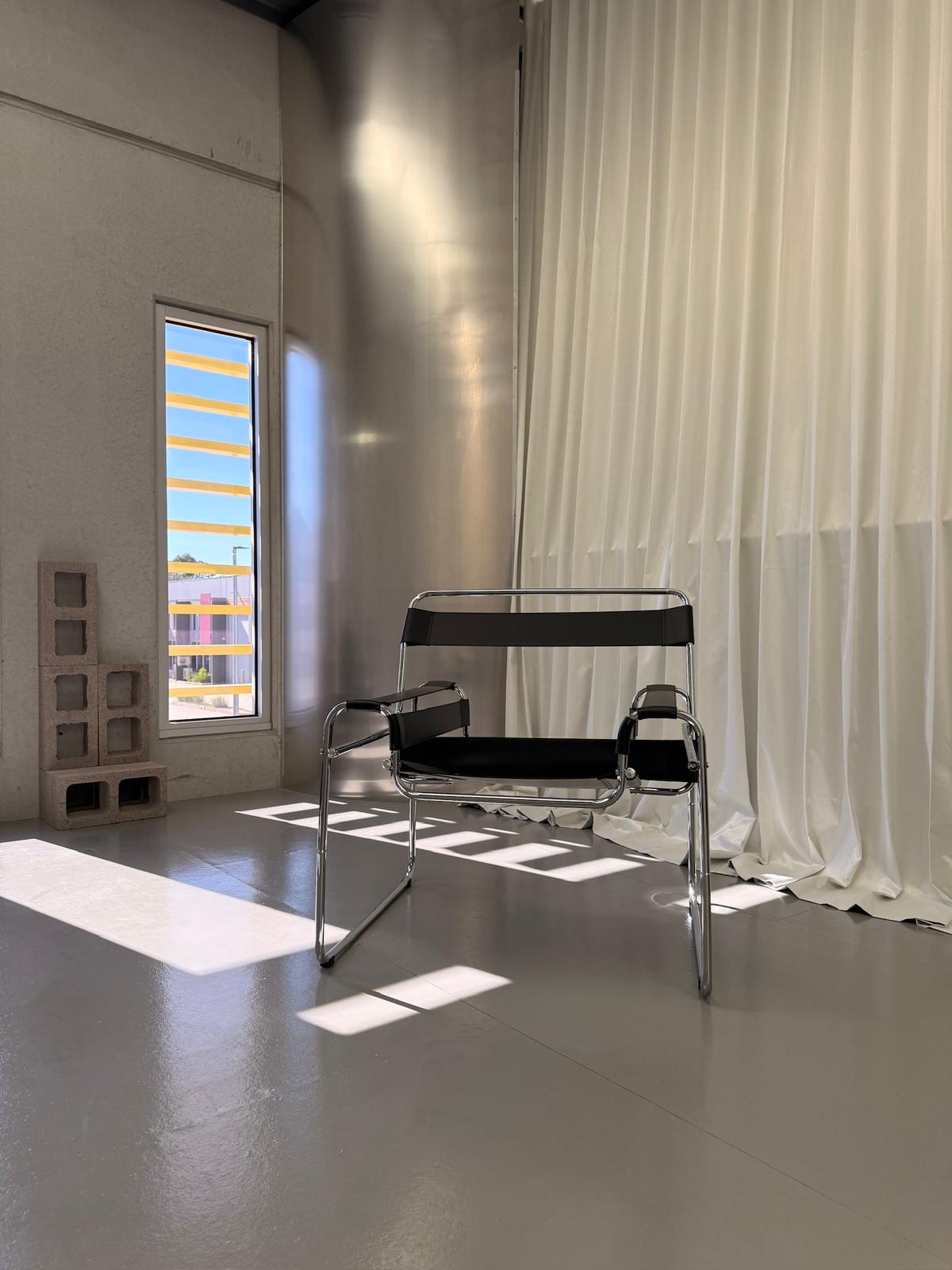 Daylight pouring through the studio's tall windows past block-out curtains, illuminating a chrome Wassily chair on polished concrete.
