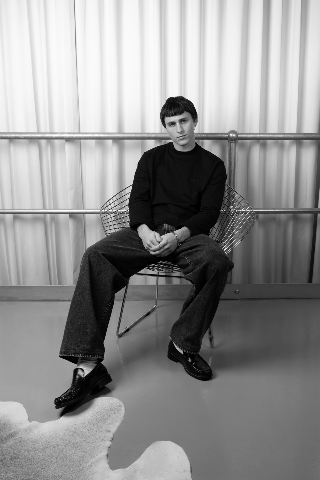Black and white portrait of a figure seated on the chrome wire Bertoia chair beside a sheepskin rug.
