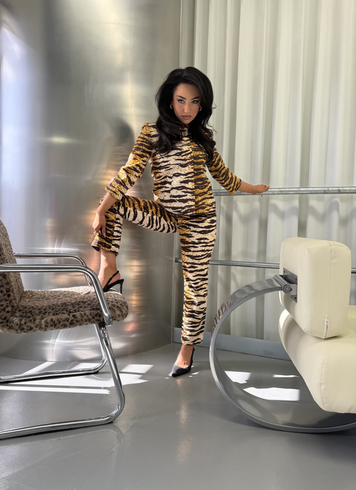A model in a tiger-print set stands between the chrome chairs in the light-filled studio.